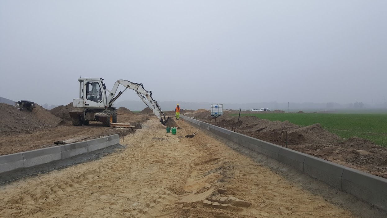 Układanie obrzeży betonowych drogi z użyciem koparki. Widoczny pracownik w odblaskowej kamizelce. Tło: pole i zamglone niebo.