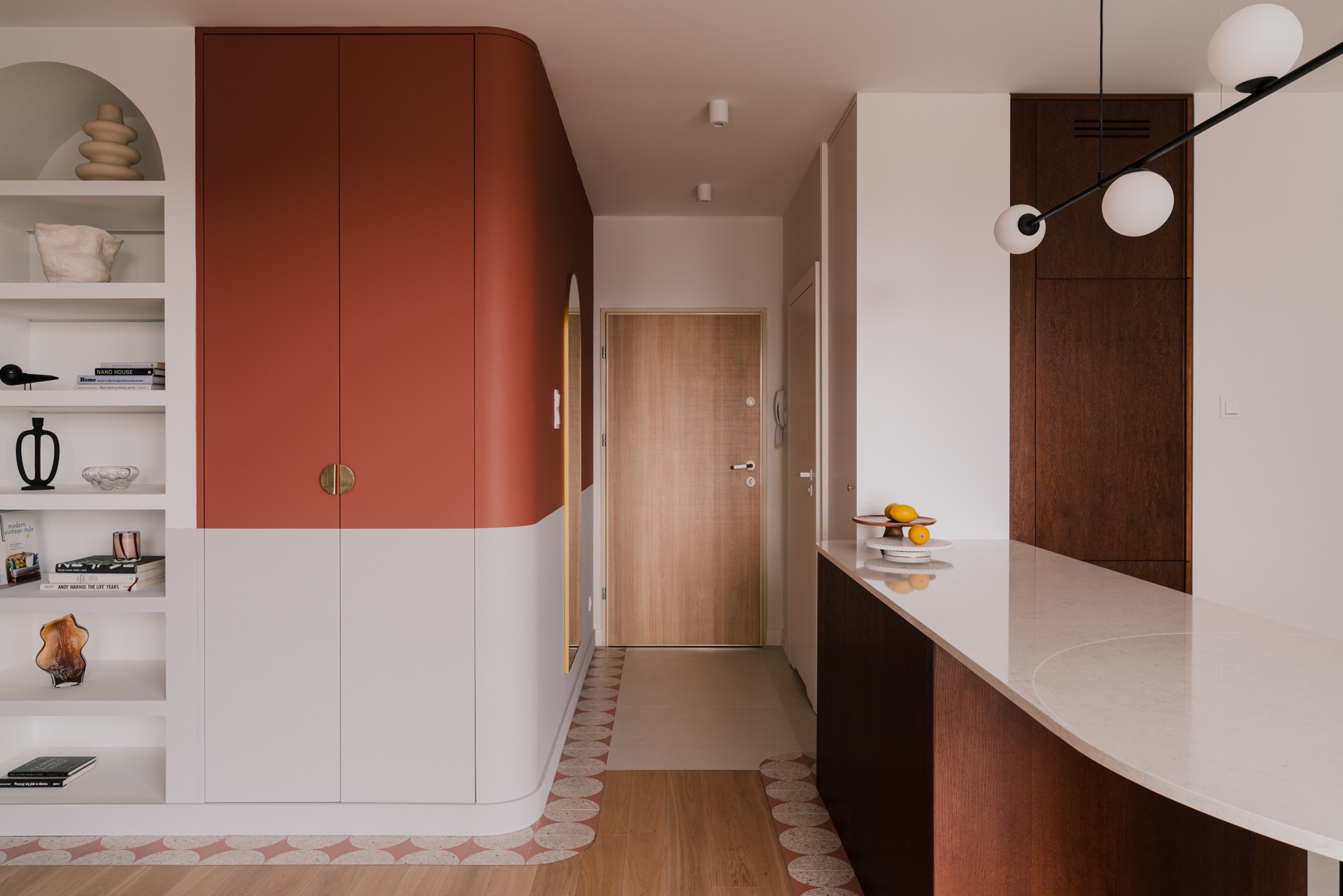 Nowoczesne wnętrze mieszkania: zabudowa z szafkami w kolorze terakoty i białym, regał z dekoracjami, zaokrąglony blat i dekoracyjna podłoga z motywem kół.