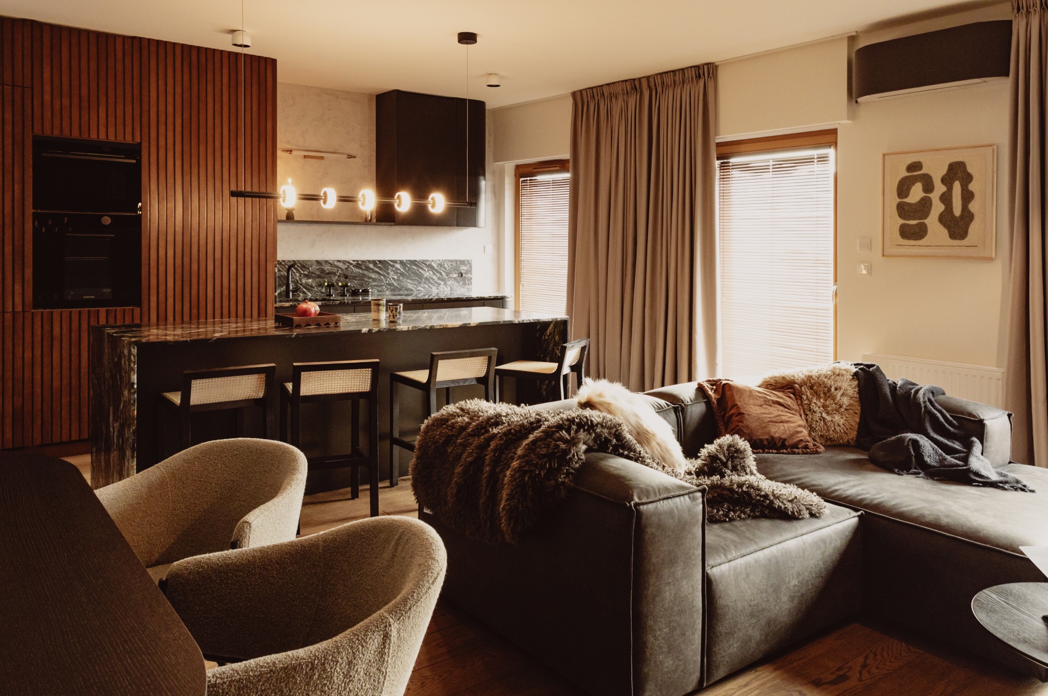 Ciepłe, nowoczesne wnętrze z drewnianą ścianą, marmurową wyspą kuchenną i wygodnym narożnikiem z poduszkami. Subtelne oświetlenie i naturalne materiały.