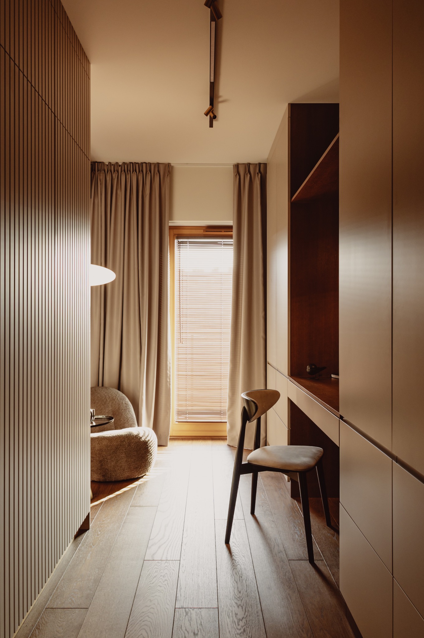 Neutralna kolorystyka gabinetu: drewniane panele ścienne, biurko z szafkami, krzesło i fotel boucle. Okno z żaluzjami i zasłonami w tle. Ciepłe, naturalne światło.