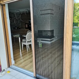 INTEGRO POZNAŃ SPÓŁKA Z OGRANICZONĄ ODPOWIEDZIALNOŚCIĄ - Plisowana moskitiera w drewnianej ramie, zamontowana w drzwiach tarasowych. Widok na wnętrze domu i ogród. Zabezpieczenie przed owadami w nowoczesnym wydaniu.