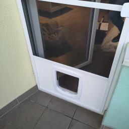 INTEGRO POZNAŃ SPÓŁKA Z OGRANICZONĄ ODPOWIEDZIALNOŚCIĄ - Biała moskitiera z klapką dla zwierząt, zamontowana w drzwiach balkonowych. Widoczny fragment wnętrza z kwiatami i poduszką na podłodze. U dołu szare płytki balkonowe.