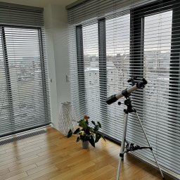 INTEGRO POZNAŃ SPÓŁKA Z OGRANICZONĄ ODPOWIEDZIALNOŚCIĄ - Jasne, poziome żaluzje w nowoczesnym pokoju z teleskopem. Widok na miasto przez okno. Drewniana podłoga, roślina w doniczce i dekoracyjny lampion.