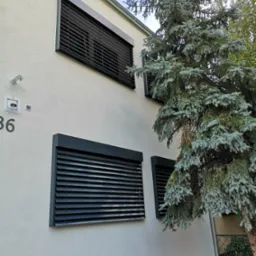 Elewacja budynku z zamontowanymi, ciemnymi żaluzjami fasadowymi, częściowo zasłoniętymi przez iglaste drzewo. Widoczny numer domu i fragment rynny.