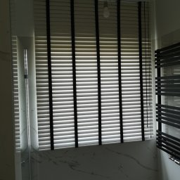 INTEGRO POZNAŃ SPÓŁKA Z OGRANICZONĄ ODPOWIEDZIALNOŚCIĄ - Zamknięte poziome żaluzje w łazience z marmurowym wykończeniem, widoczne elementy kabiny prysznicowej i grzejnika drabinkowego. Oświetlenie sufitowe w tle.