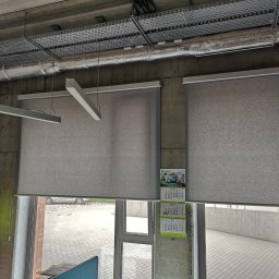 INTEGRO POZNAŃ SPÓŁKA Z OGRANICZONĄ ODPOWIEDZIALNOŚCIĄ - Dwie szare rolety wewnętrzne zamontowane w oknach. Widoczny fragment sufitu z instalacjami. Na ścianie kalendarz firmowy. Za oknem betonowy mur i fragment parkingu.