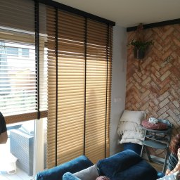 INTEGRO POZNAŃ SPÓŁKA Z OGRANICZONĄ ODPOWIEDZIALNOŚCIĄ - Beżowe żaluzje drewniane z czarnymi elementami, zamontowane w oknie w salonie z ceglaną ścianą i niebieską sofą. Widoczne detale wystroju wnętrza.