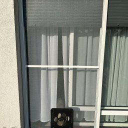 INTEGRO POZNAŃ SPÓŁKA Z OGRANICZONĄ ODPOWIEDZIALNOŚCIĄ - Biała moskitiera z otworem dla zwierząt domowych w oknie, z widoczną zasłoną w tle i fragmentem rolety zewnętrznej po prawej stronie.