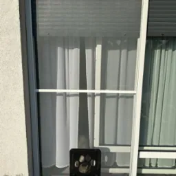 Biała moskitiera z otworem dla zwierząt domowych w oknie, z widoczną zasłoną w tle i fragmentem rolety zewnętrznej po prawej stronie.