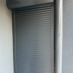 Zamknięta, grafitowa roleta zewnętrzna z widoczną rurą spustową na szarej, teksturowanej elewacji budynku. Widok z bliska, światło dzienne.