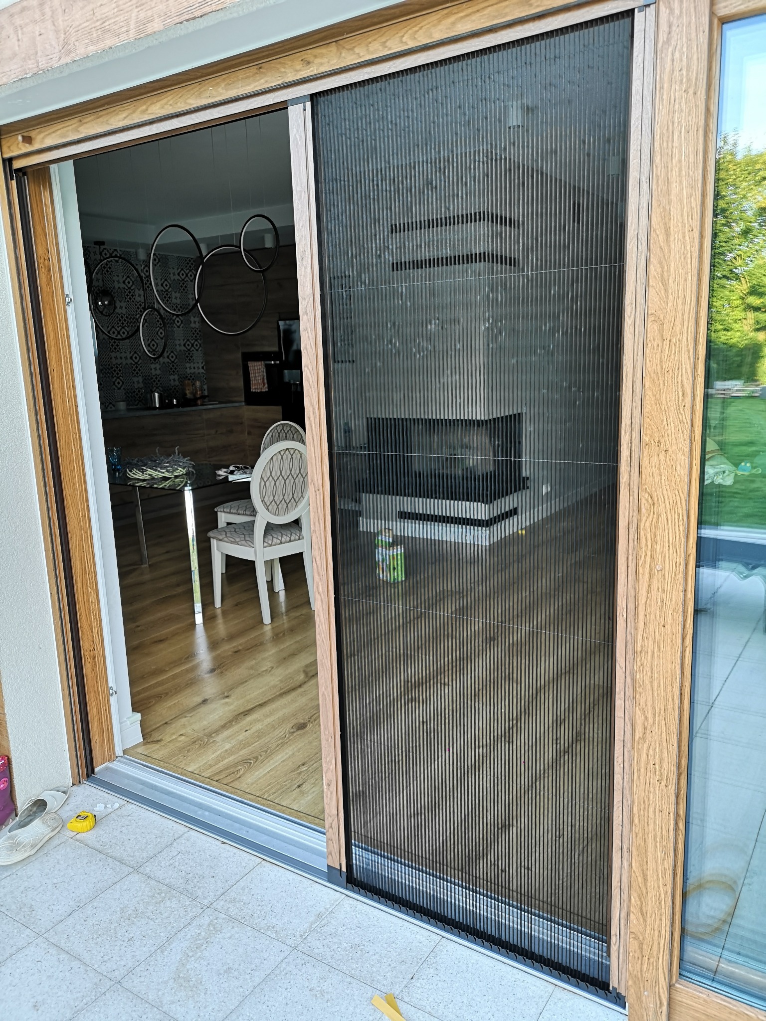 Plisowana moskitiera w drewnianej ramie, zamontowana w drzwiach tarasowych. Widok na wnętrze domu i ogród. Zabezpieczenie przed owadami w nowoczesnym wydaniu.