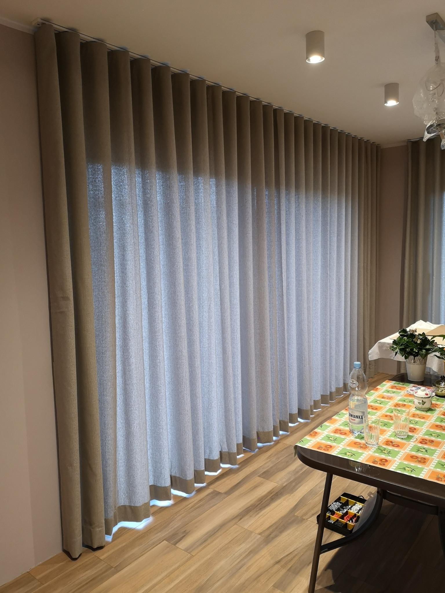 Beżowe, plisowane zasłony na szynie sufitowej, z widocznym wykończeniem na dole, w jasnym wnętrzu z drewnianą podłogą i stołem nakrytym obrusem.