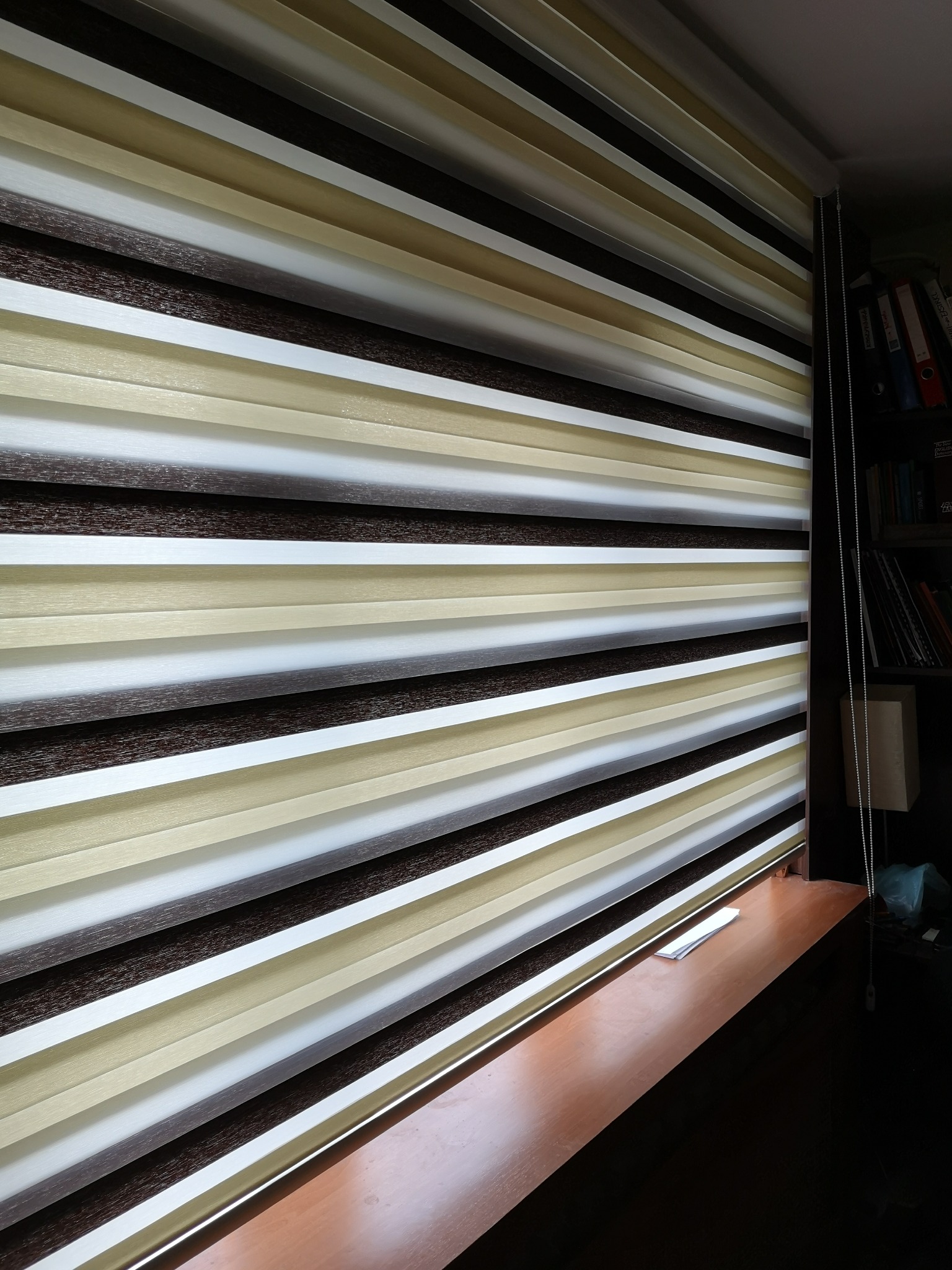 Zasłonięte okno z roletą dzień noc w odcieniach brązu, beżu i bieli, z widocznym łańcuszkiem sterującym i fragmentem półki okiennej.