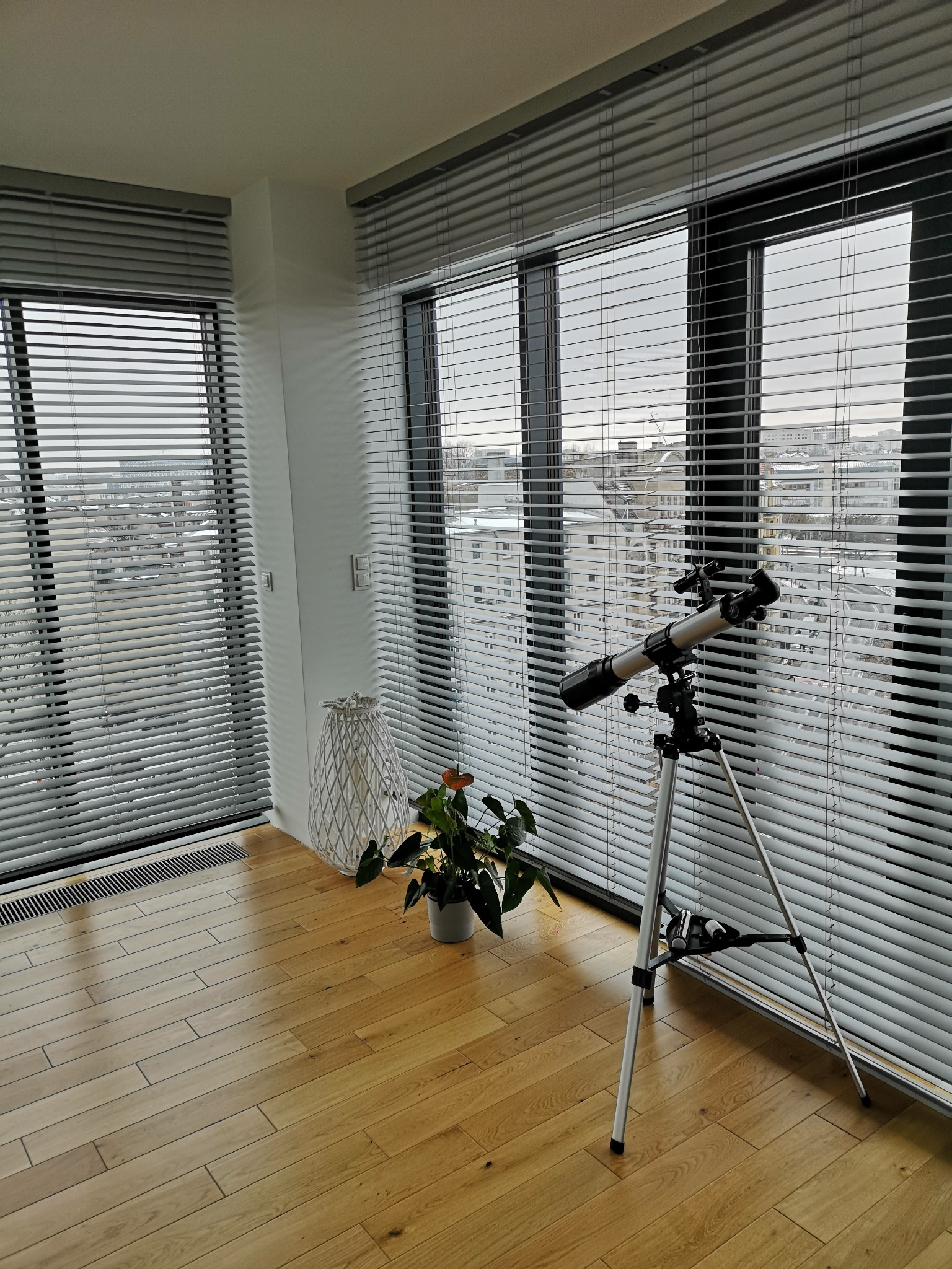 Jasne, poziome żaluzje w nowoczesnym pokoju z teleskopem. Widok na miasto przez okno. Drewniana podłoga, roślina w doniczce i dekoracyjny lampion.