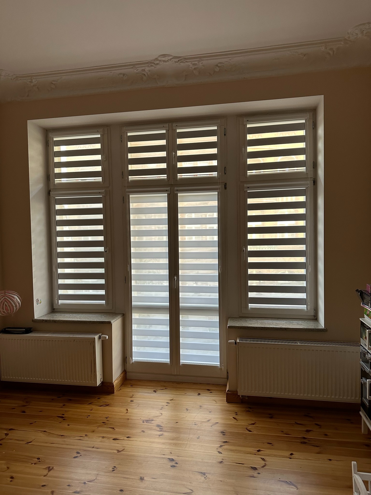 Trzyczęściowe okno z zamontowanymi roletami dzień i noc, ustawionymi w poziome pasy. Widok na drewnianą podłogę i grzejniki pod oknami. Dekoracyjny sufit.