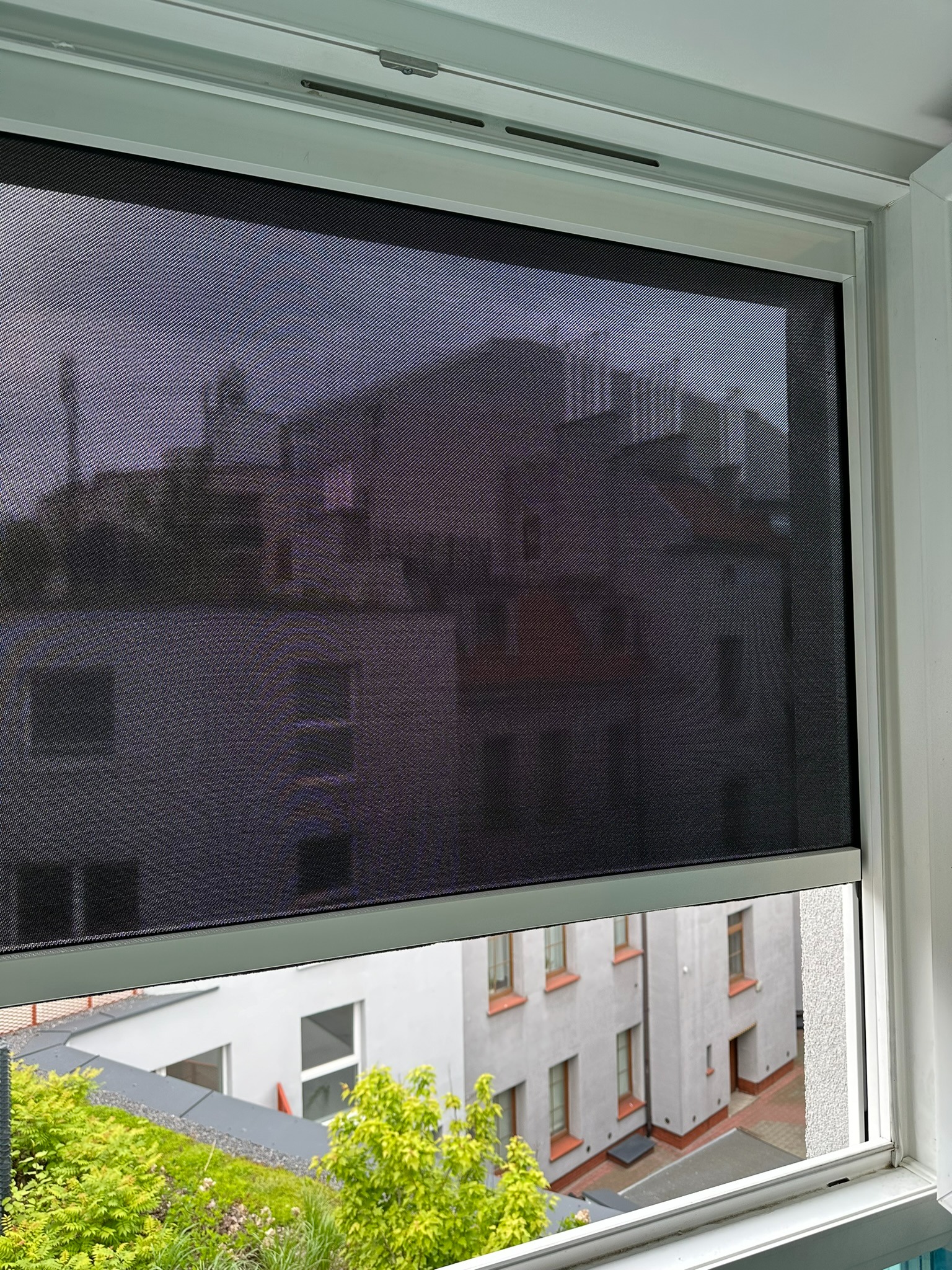 Zamknięta, grafitowa roleta wewnętrzna w białej ramie okiennej, z widokiem na zieleń i budynki na zewnątrz. Delikatna faktura materiału rolety.