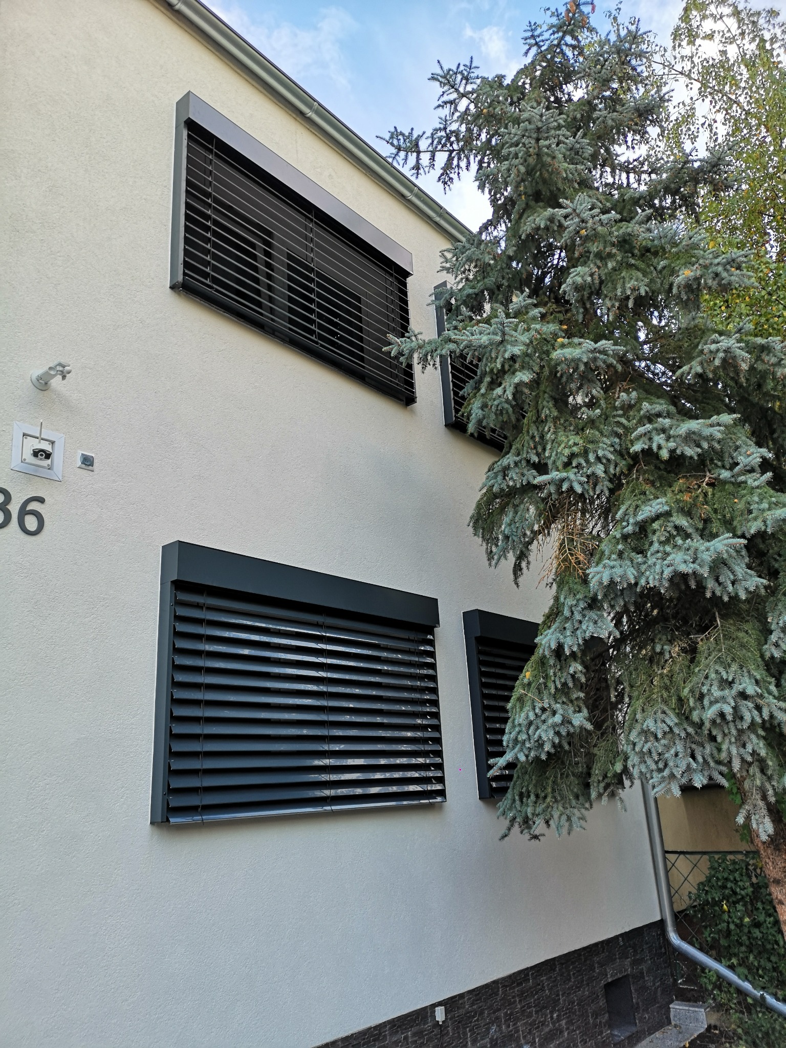 Elewacja budynku z zamontowanymi, ciemnymi żaluzjami fasadowymi, częściowo zasłoniętymi przez iglaste drzewo. Widoczny numer domu i fragment rynny.