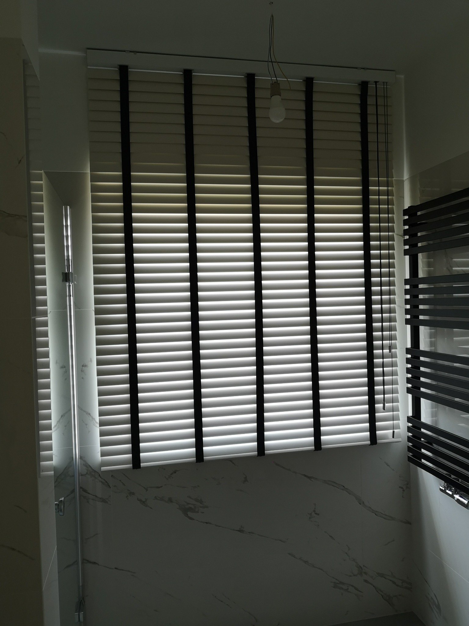 Zamknięte poziome żaluzje w łazience z marmurowym wykończeniem, widoczne elementy kabiny prysznicowej i grzejnika drabinkowego. Oświetlenie sufitowe w tle.