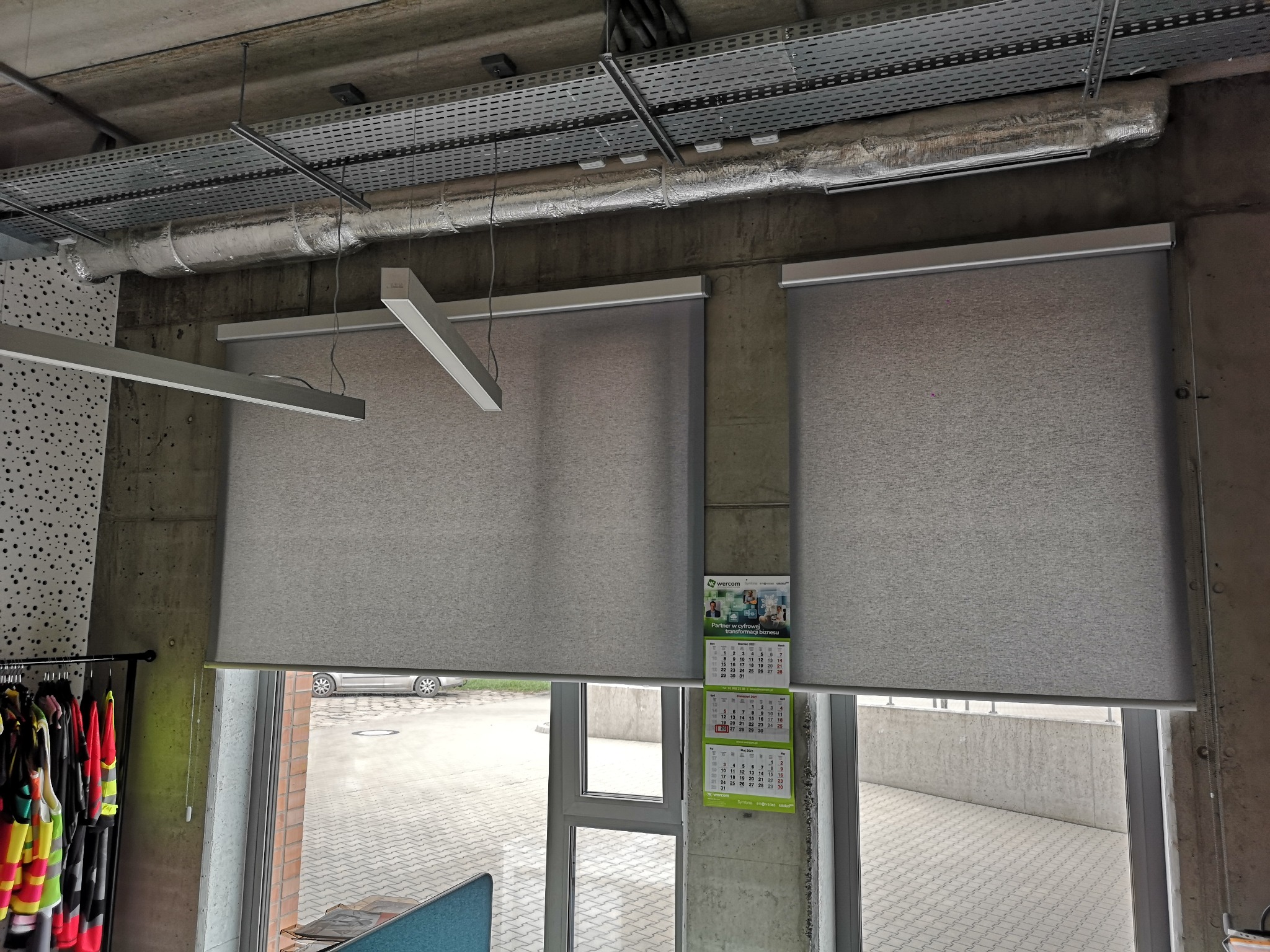 Dwie szare rolety wewnętrzne zamontowane w oknach. Widoczny fragment sufitu z instalacjami. Na ścianie kalendarz firmowy. Za oknem betonowy mur i fragment parkingu.