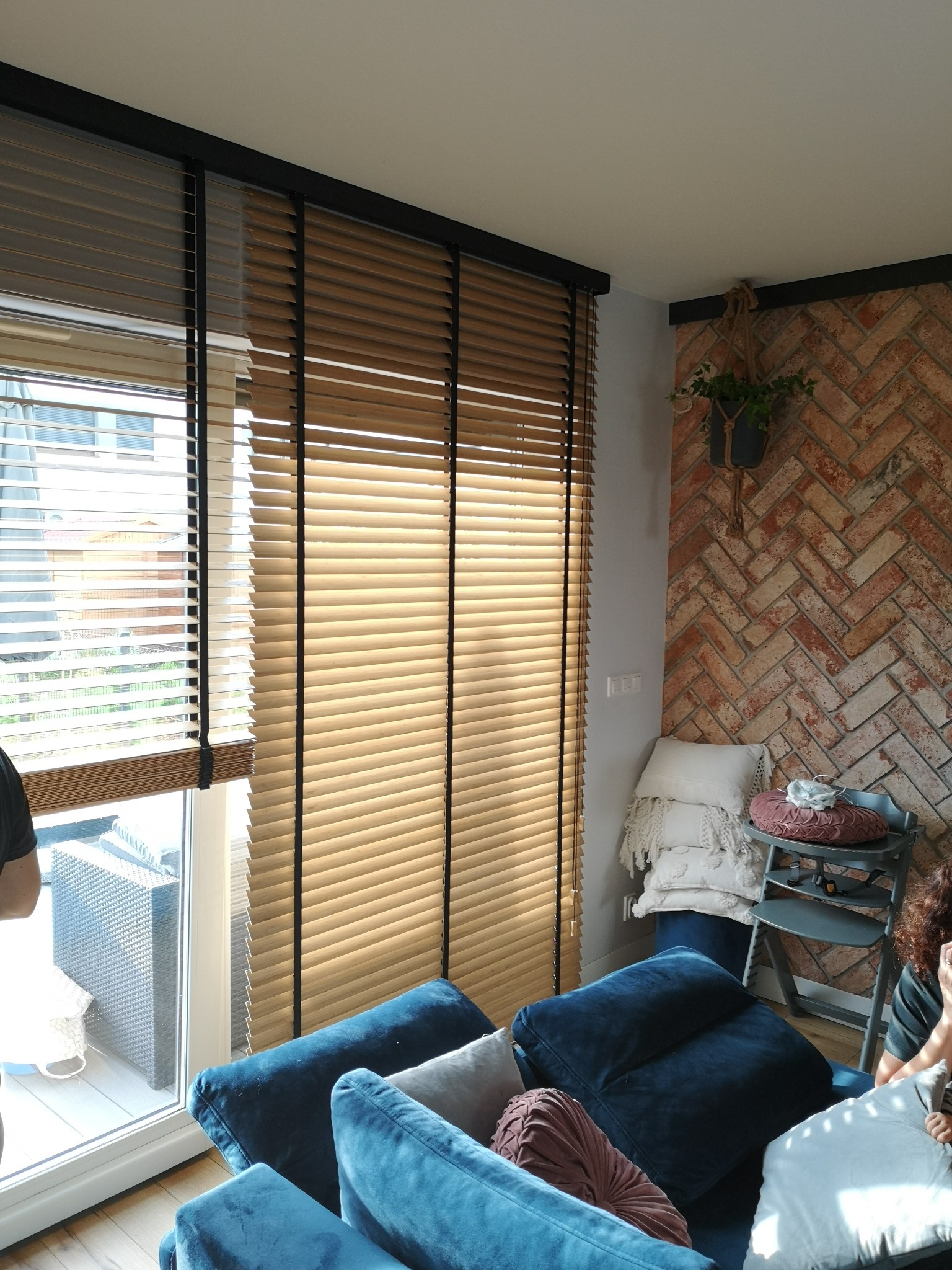 Beżowe żaluzje drewniane z czarnymi elementami, zamontowane w oknie w salonie z ceglaną ścianą i niebieską sofą. Widoczne detale wystroju wnętrza.