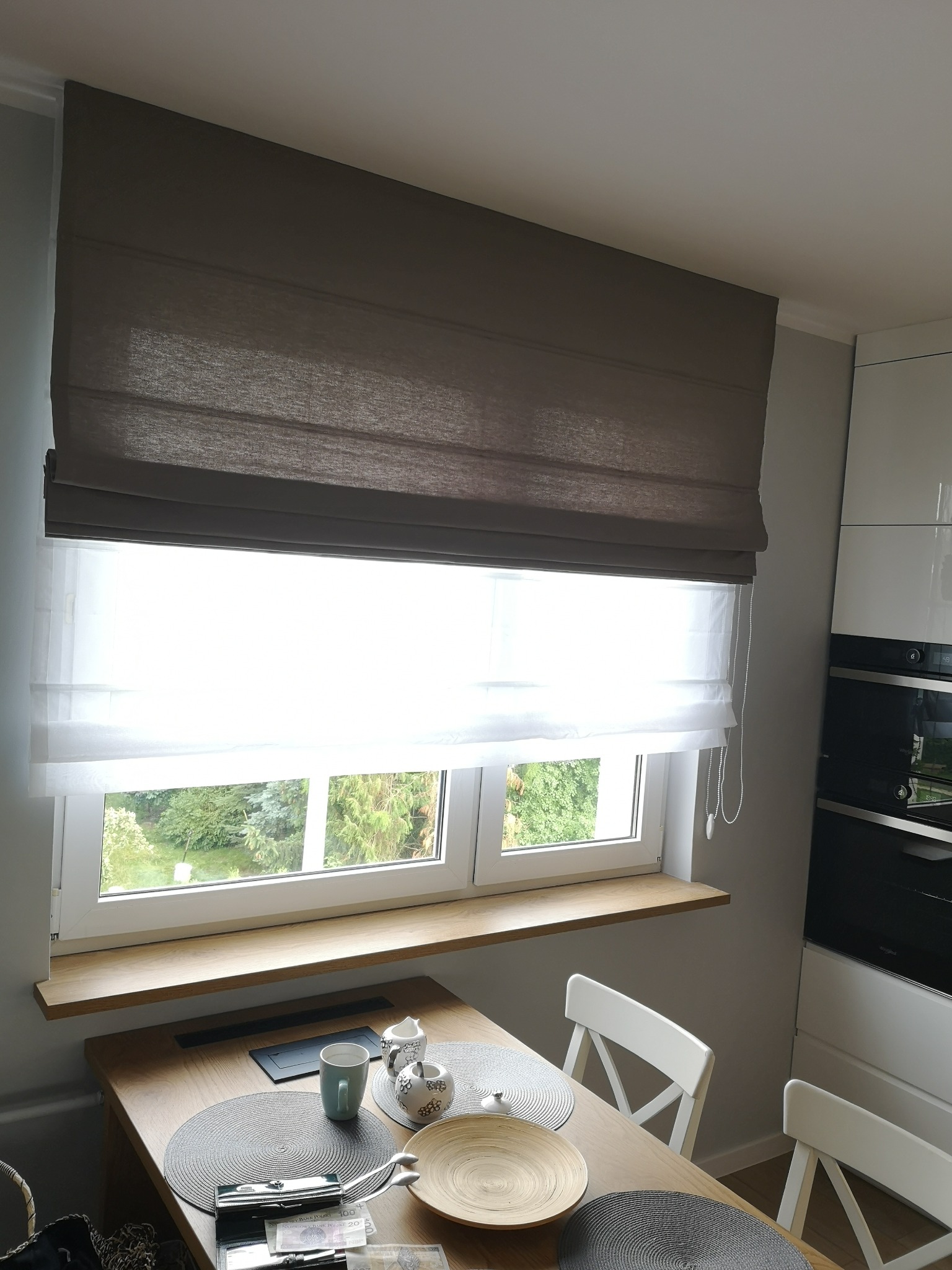 Beżowa roleta rzymska z białym woalem w oknie. Na parapecie drewniany blat. Widok na zieleń za oknem. Ustawiony stół z nakryciem. Nowoczesna kuchnia w tle.