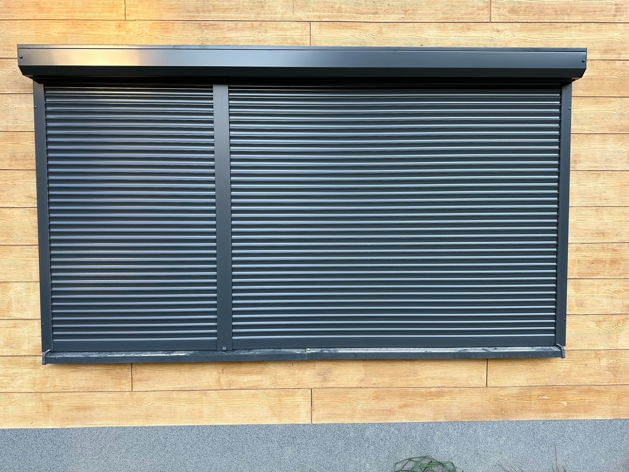 Zamknięta, grafitowa roleta zewnętrzna na tle drewnianej elewacji. Widoczne poziome lamele i kaseta rolety. Całość w naturalnym oświetleniu.