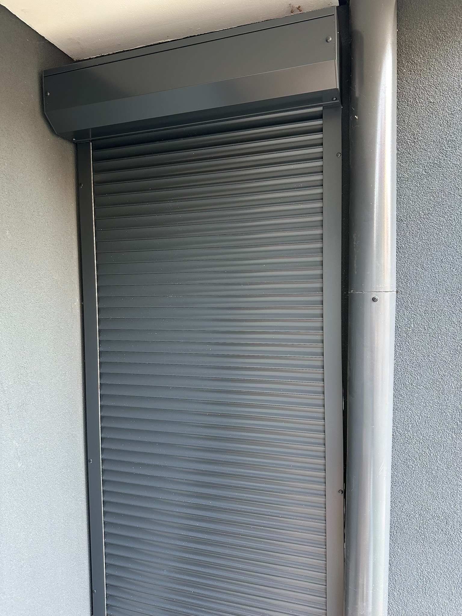 Zamknięta, grafitowa roleta zewnętrzna z widoczną rurą spustową na szarej, teksturowanej elewacji budynku. Widok z bliska, światło dzienne.