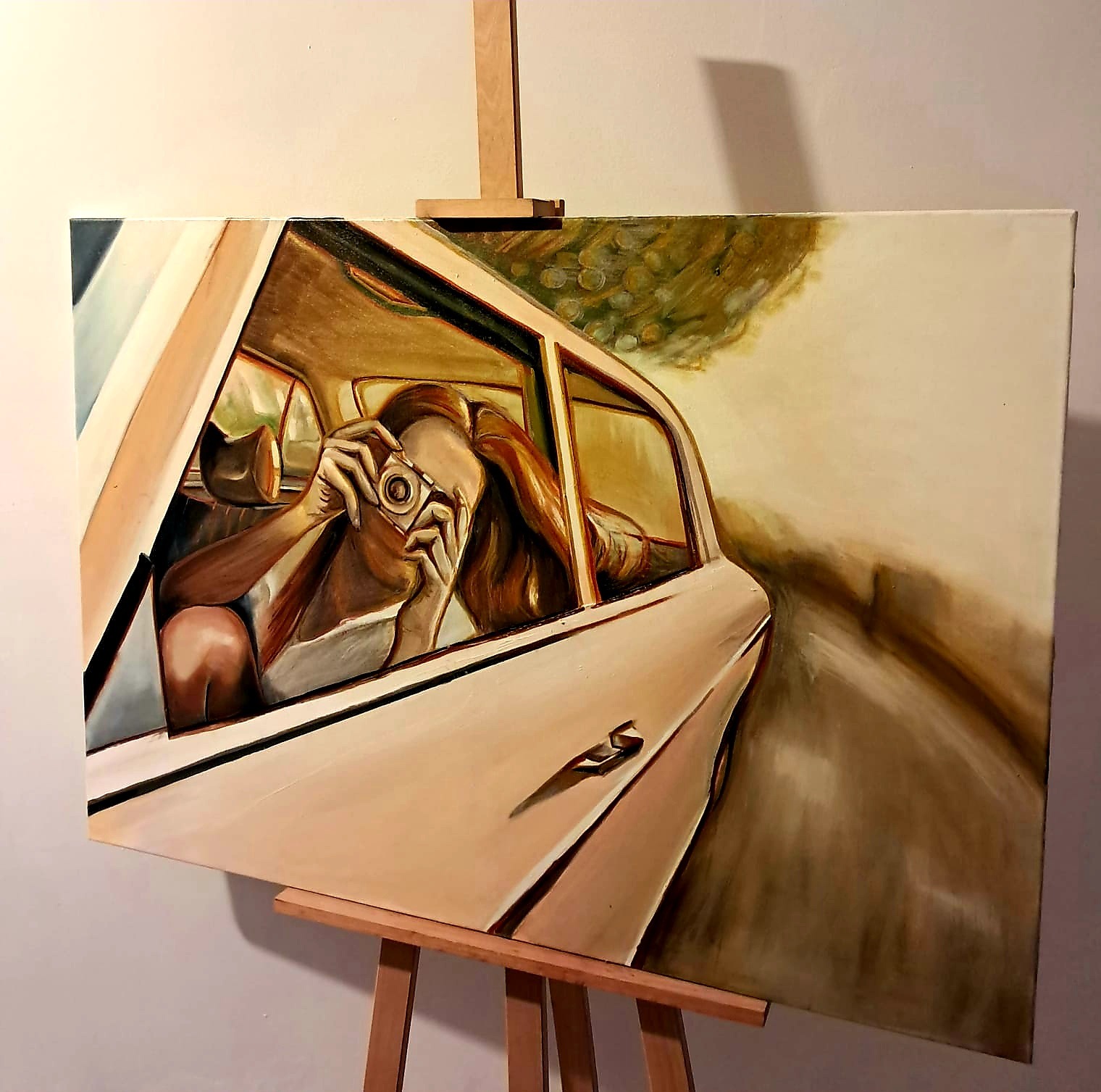 Obraz olejny na płótnie przedstawiający kobietę w samochodzie robiącą zdjęcie aparatem. Płótno umieszczone na drewnianej sztaludze, jasne tło.