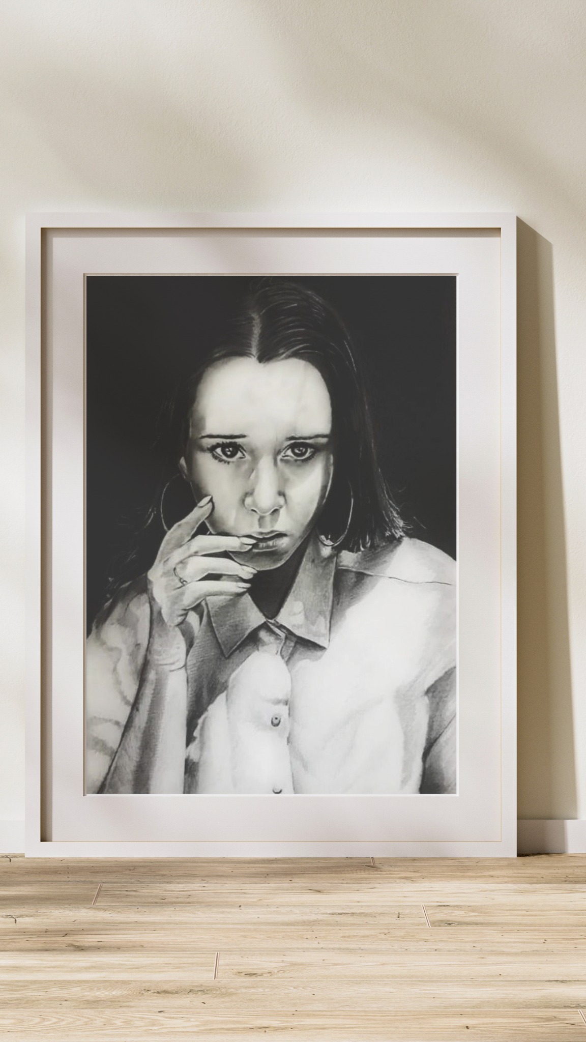 Portret kobiety w koszuli, wykonany ołówkiem i oprawiony w białą ramę, stojący na podłodze z paneli. Realistyczny rysunek z detalami.