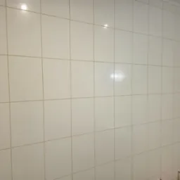 Ściana w łazience wyłożona jasnymi, kwadratowymi płytkami z widocznymi fugami. W prawym górnym rogu widoczny fragment listwy sufitowej. Minimalistyczny design.