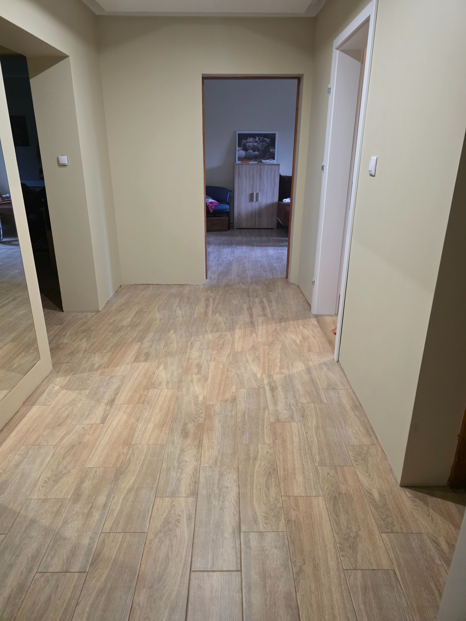 Jasne, drewnopodobne płytki ułożone w korytarzu. Widok na wnętrze mieszkania przez otwarte drzwi. Neutralne ściany i białe drzwi w tle.