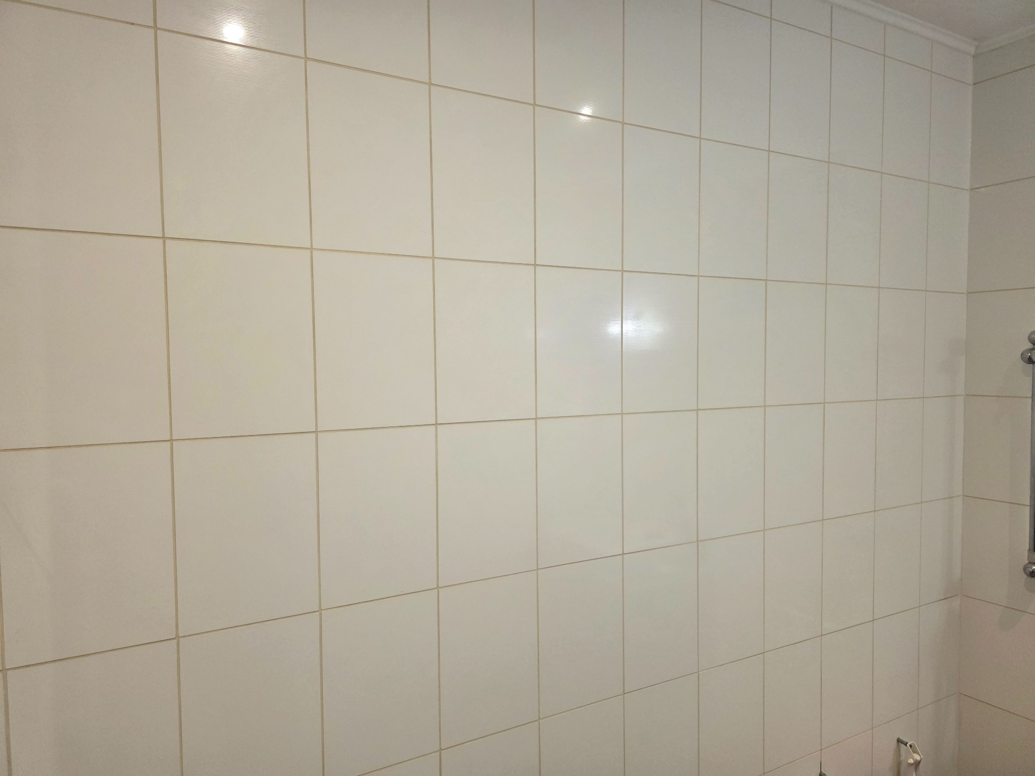 Ściana w łazience wyłożona jasnymi, kwadratowymi płytkami z widocznymi fugami. W prawym górnym rogu widoczny fragment listwy sufitowej. Minimalistyczny design.