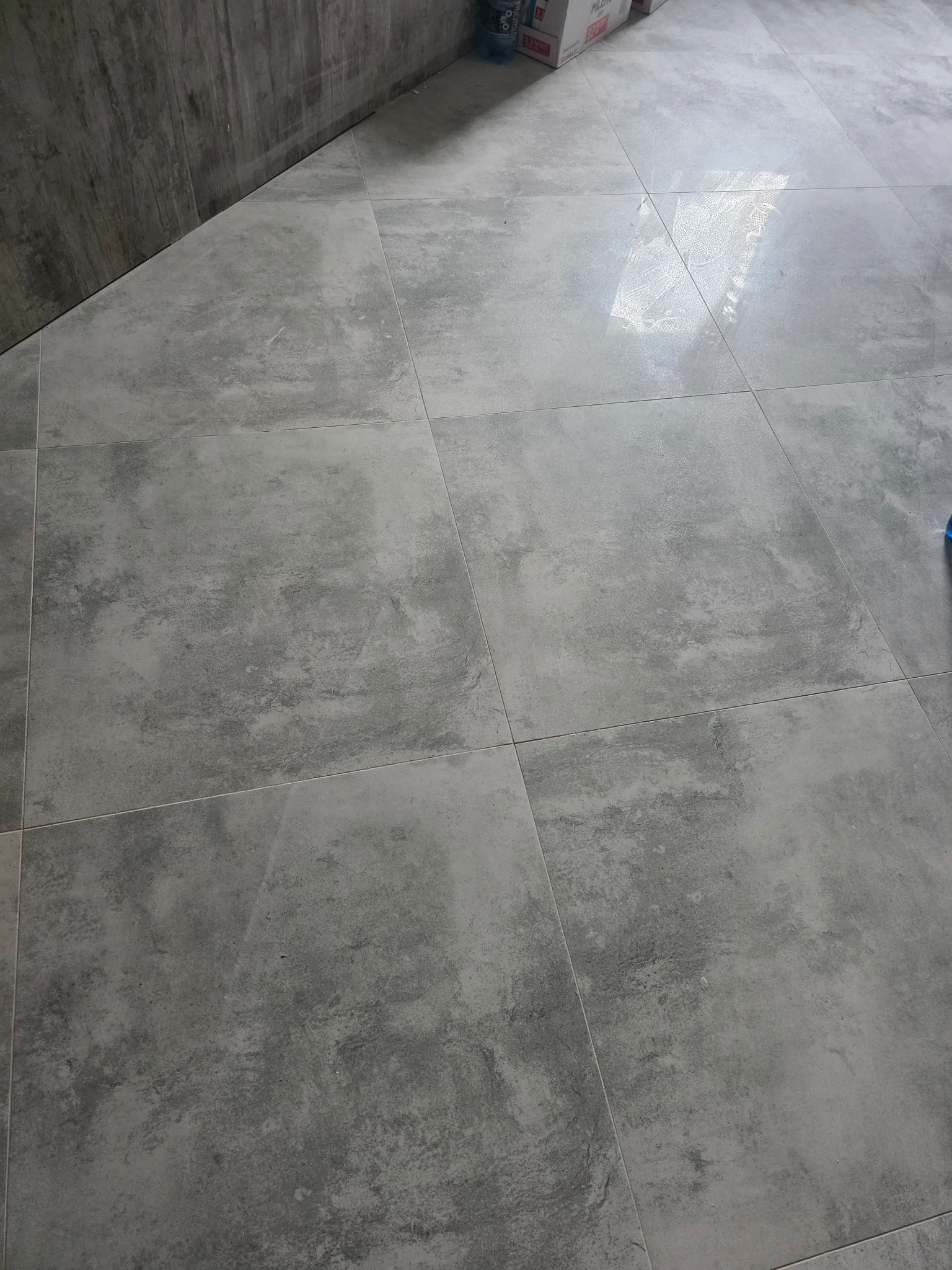 Szare, matowe płytki podłogowe ułożone w kwadraty, widoczne spoiny. Częściowo widoczna ściana i kartony w rogu. Podłoga po remoncie.