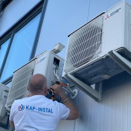 KAP-Instal - Monter instaluje klimatyzator Mitsubishi Electric na elewacji budynku. Widoczne narzędzia, jednostki zewnętrzne i logo firmy instalacyjnej na koszulce montera.
