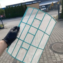 KAP-Instal - Czysty filtr klimatyzacji, trzymany w rękawicy ochronnej na zewnątrz, z napisem 'Po' w lewym górnym rogu. Widoczne budynki w tle.