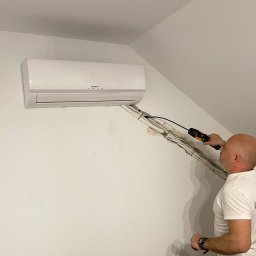 KAP-Instal - Montera klimatyzacji Kaisai sprawdza instalację elektryczną miernikiem, na białej ścianie poddasza widoczne kable i klimatyzator.