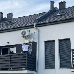 KAP-Instal - Montaż klimatyzacji na elewacji budynku. Mężczyzna na drabinie instaluje jednostkę zewnętrzną klimatyzatora. Widoczne okna z roletami i elementy dachu.