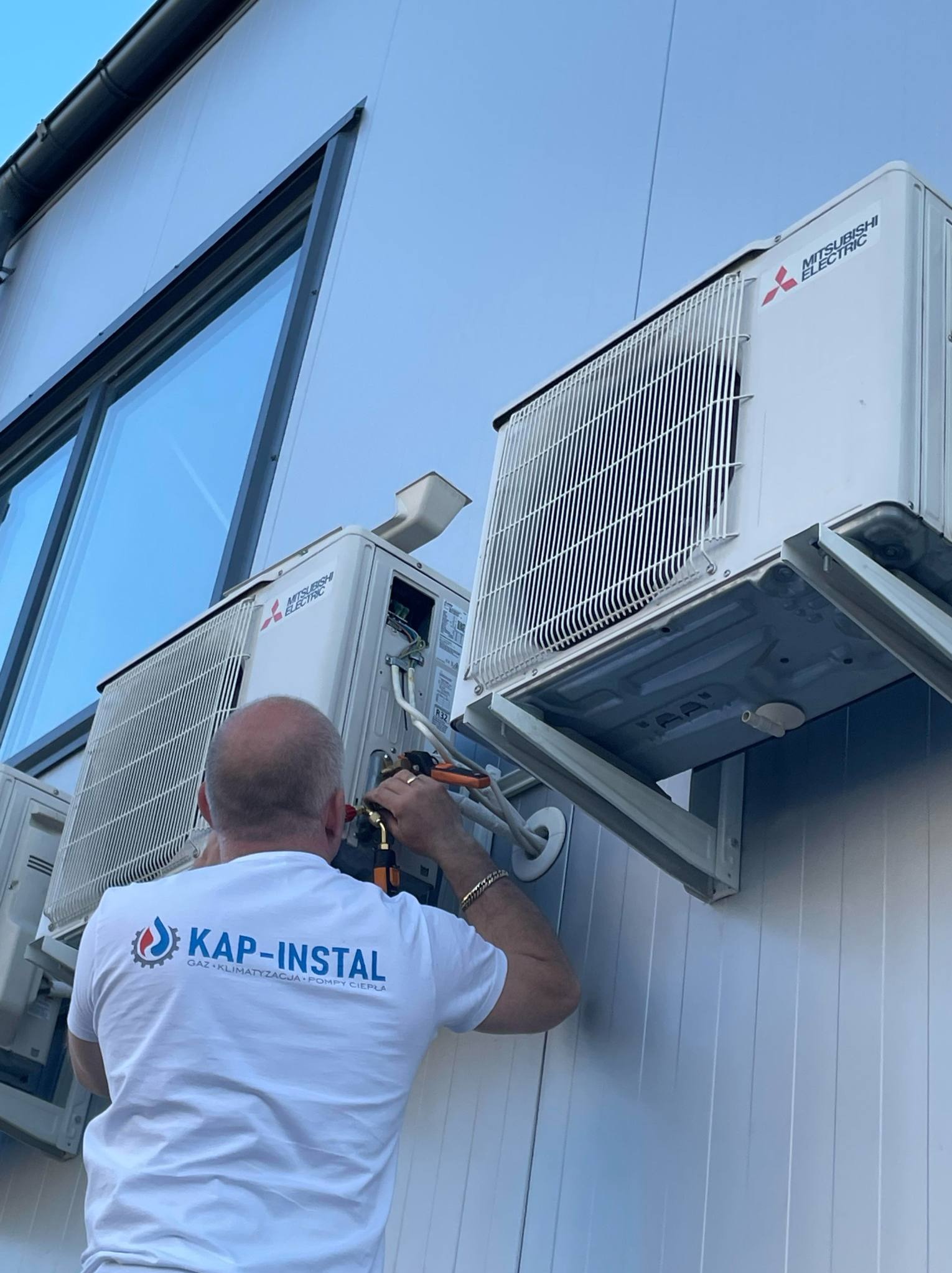 Monter instaluje klimatyzator Mitsubishi Electric na elewacji budynku. Widoczne narzędzia, jednostki zewnętrzne i logo firmy instalacyjnej na koszulce montera.