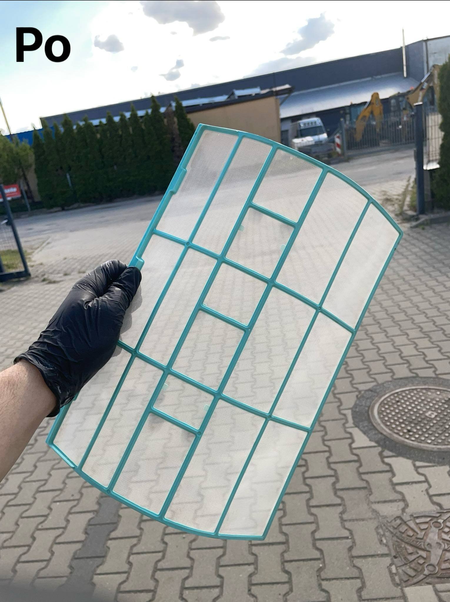 Czysty filtr klimatyzacji, trzymany w rękawicy ochronnej na zewnątrz, z napisem 'Po' w lewym górnym rogu. Widoczne budynki w tle.