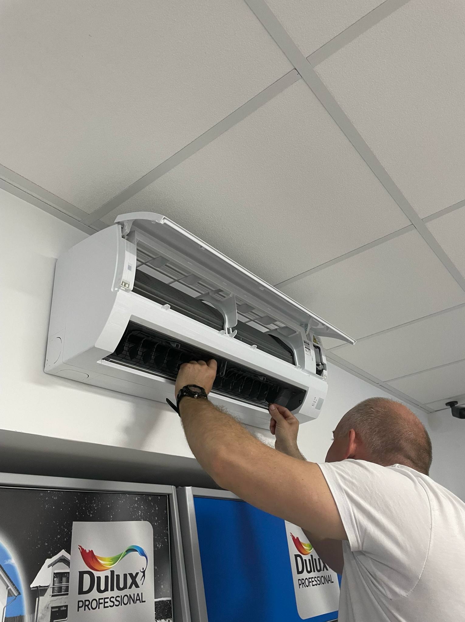 Serwisant w trakcie czyszczenia filtra klimatyzacji naściennej w pomieszczeniu biurowym. Widoczne reklamy farb Dulux Professional.