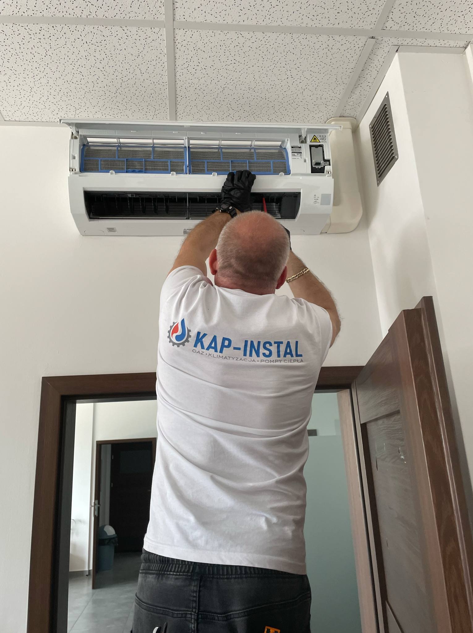 Serwisant w czarnych rękawicach sprawdza filtr klimatyzacji w pomieszczeniu z białym sufitem kasetonowym, widoczny fragment logotypu firmy na koszulce.