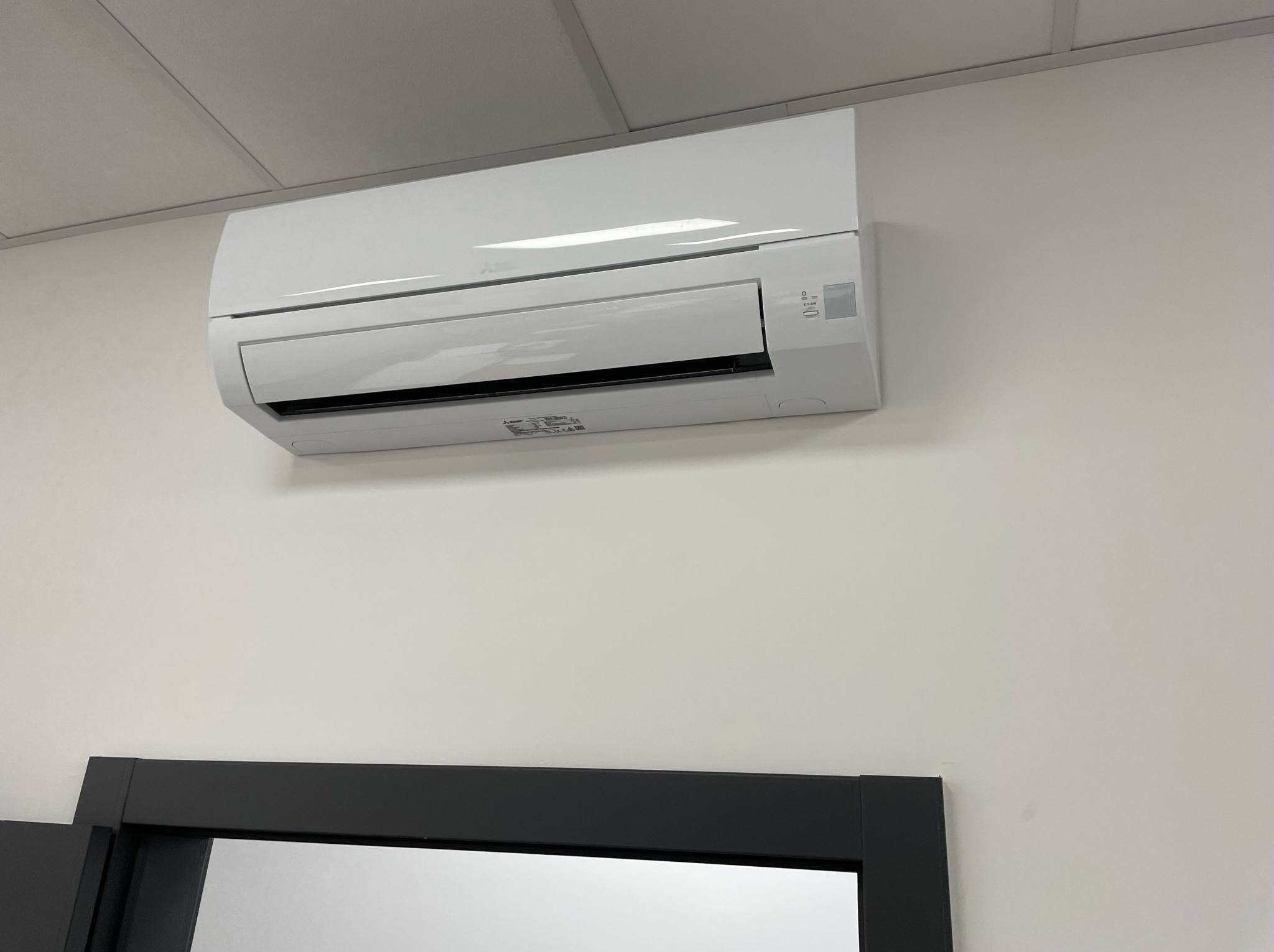 Biała klimatyzacja Mitsubishi zamontowana na ścianie nad czarną futryną drzwi. Widok z dołu, oświetlenie dzienne.