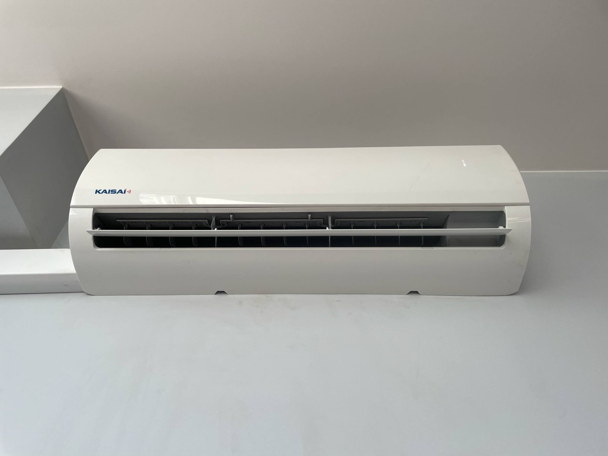 Biały klimatyzator Kaisai zamontowany na ścianie, widok z przodu, wnętrze pomieszczenia z białymi ścianami i sufitem.