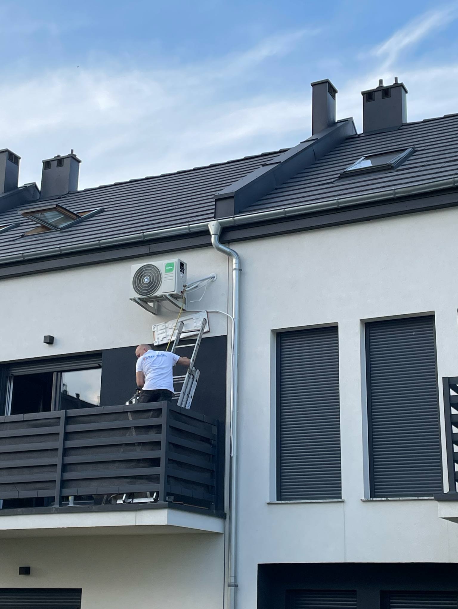 Montaż klimatyzacji na elewacji budynku. Mężczyzna na drabinie instaluje jednostkę zewnętrzną klimatyzatora. Widoczne okna z roletami i elementy dachu.
