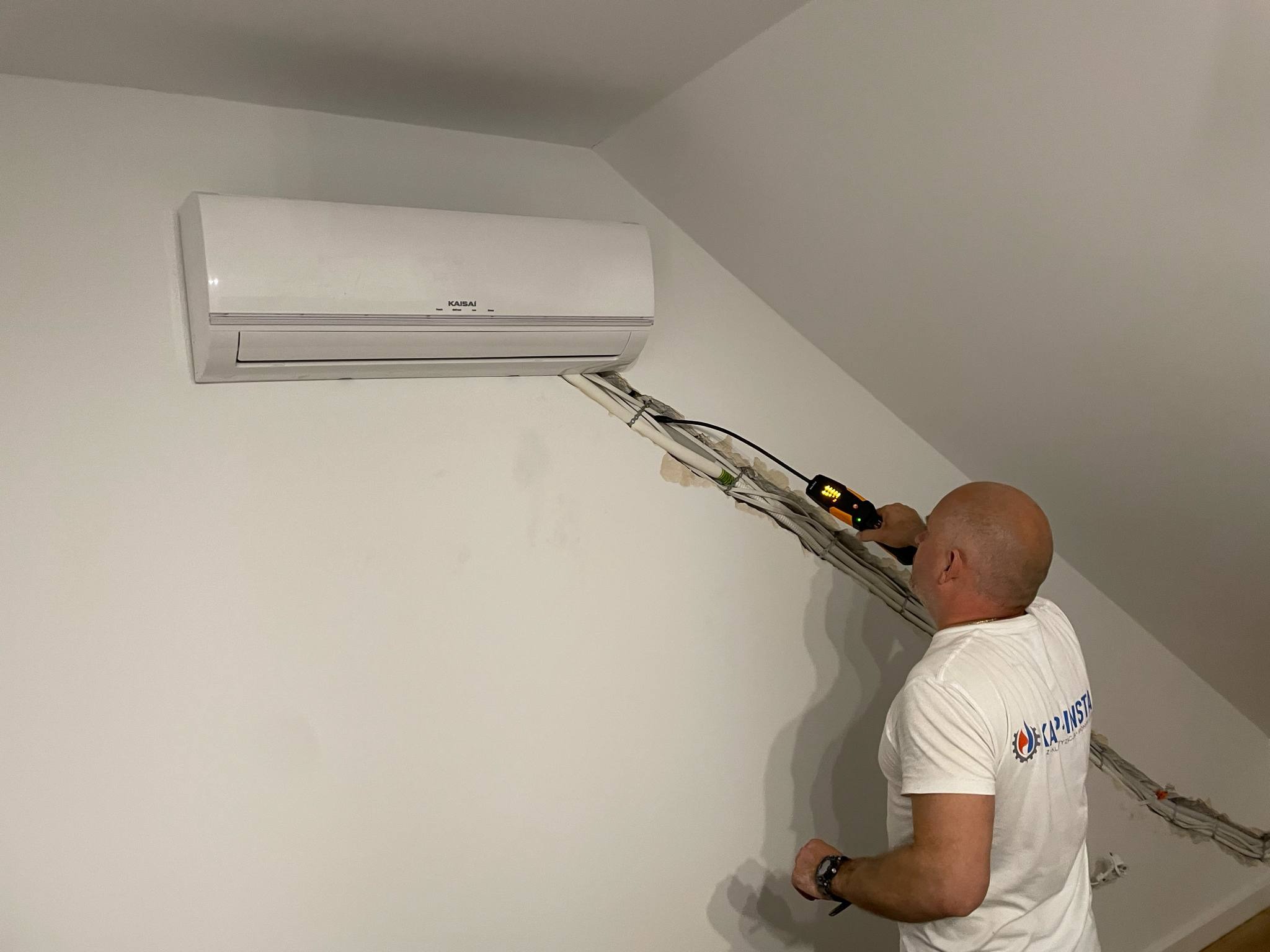 Biała klimatyzacja Kaisai zamontowana na skośnej ścianie poddasza. Instalator sprawdza kable wychodzące ze ściany za pomocą miernika.