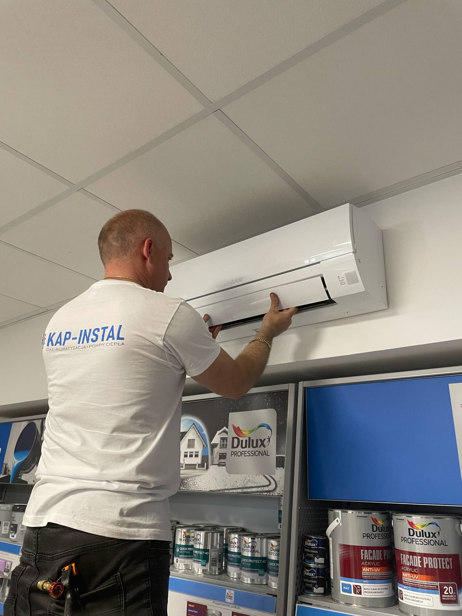Instalator w koszulce z logo firmy montuje klimatyzator Mitsubishi na ścianie. W tle widoczne farby Dulux na półkach sklepowych.