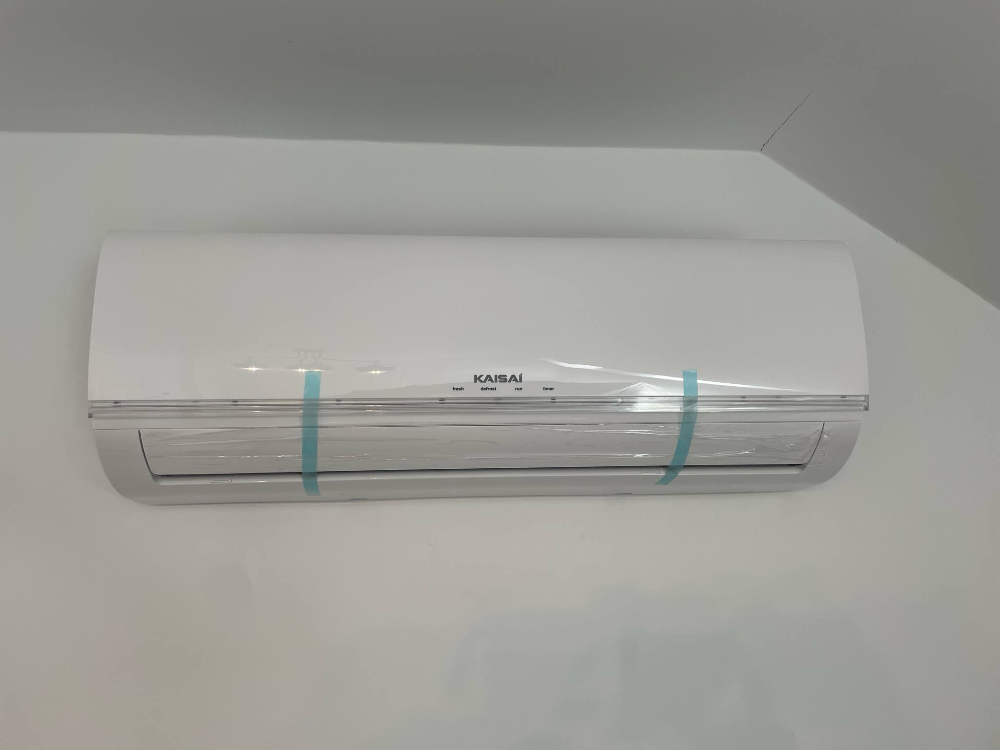 Biała klimatyzacja Kaisai, zamontowana na jasnej ścianie, zabezpieczona folią ochronną. Widoczne diody świecące. Ujęcie z bliska, z lekkim kątem.