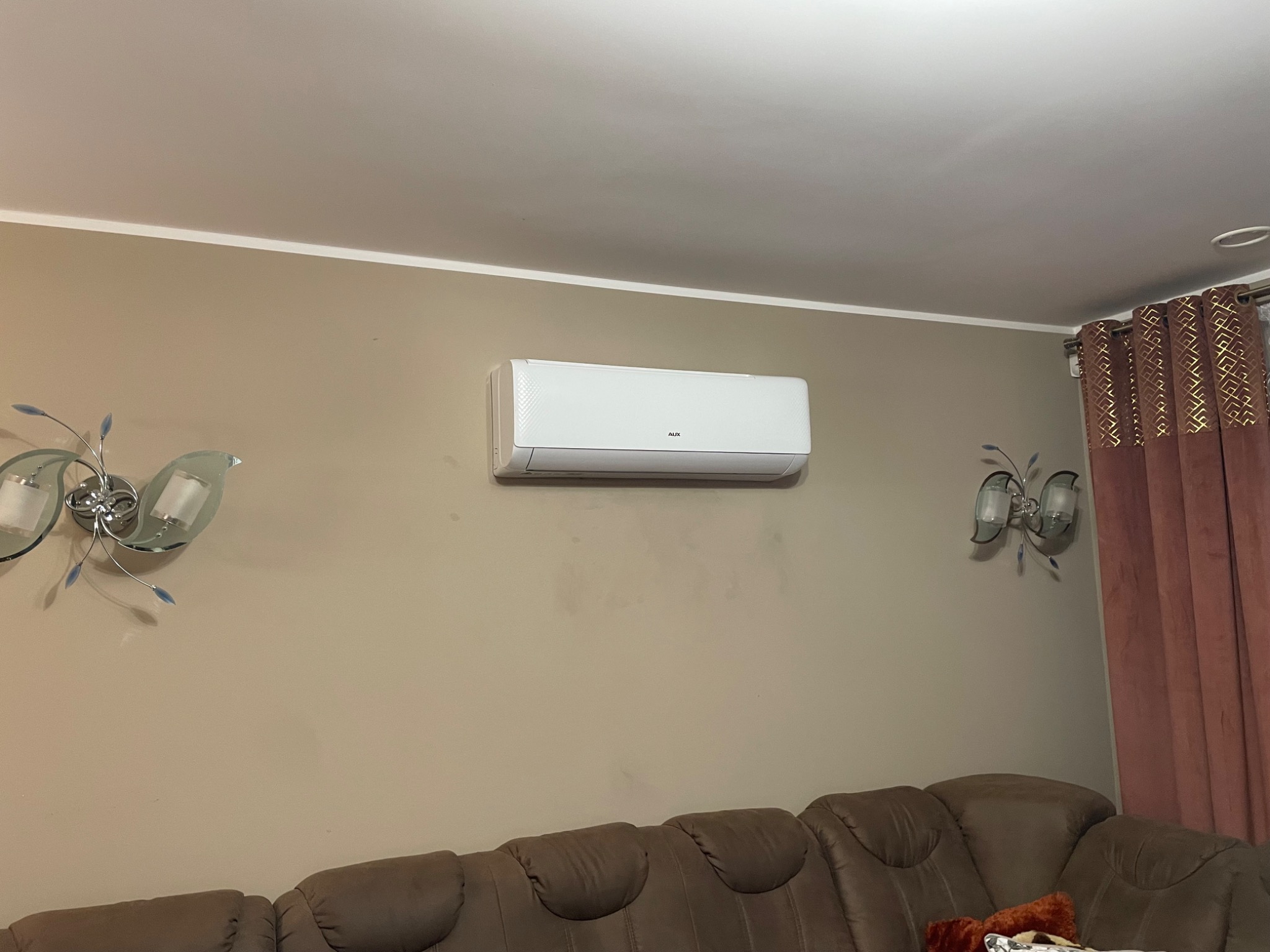 Biały klimatyzator AUX na beżowej ścianie w salonie, nad brązową sofą. Dekoracyjne lampy ścienne po bokach, zasłony w tle.