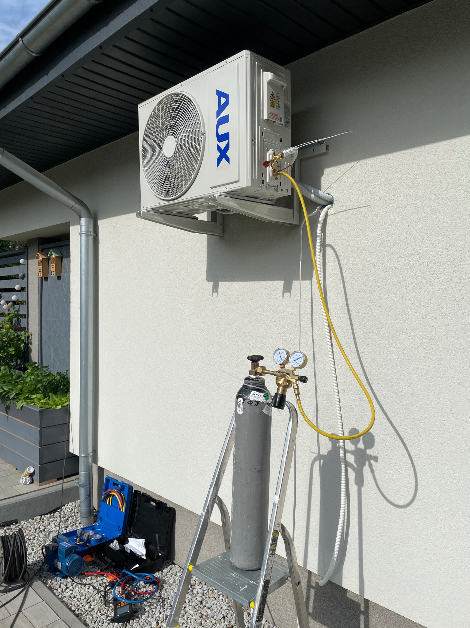 Biały klimatyzator AUX zamontowany na elewacji domu, podłączony do butli z gazem i manometrami podczas serwisu. Widoczny sprzęt serwisowy i drabina.