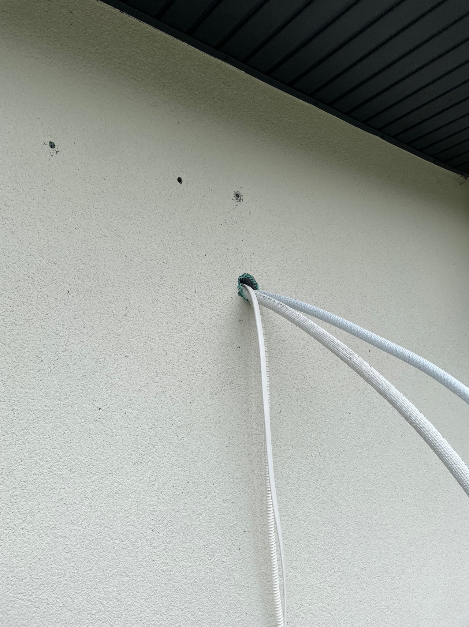 Przewody klimatyzacyjne wychodzące przez otwór w elewacji budynku. Widoczne ślady po wierceniu. Instalacja w trakcie montażu. Biała ściana i fragment podbitki.
